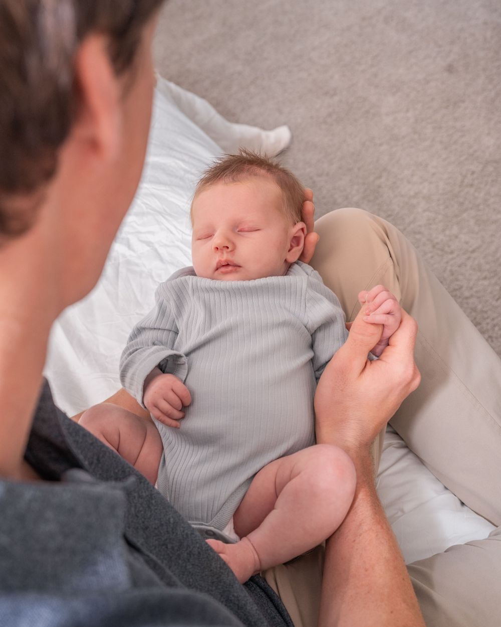 A peaceful baby sleeps in a gray onesie while being tenderly held by somebody wearing dark gray clothing.