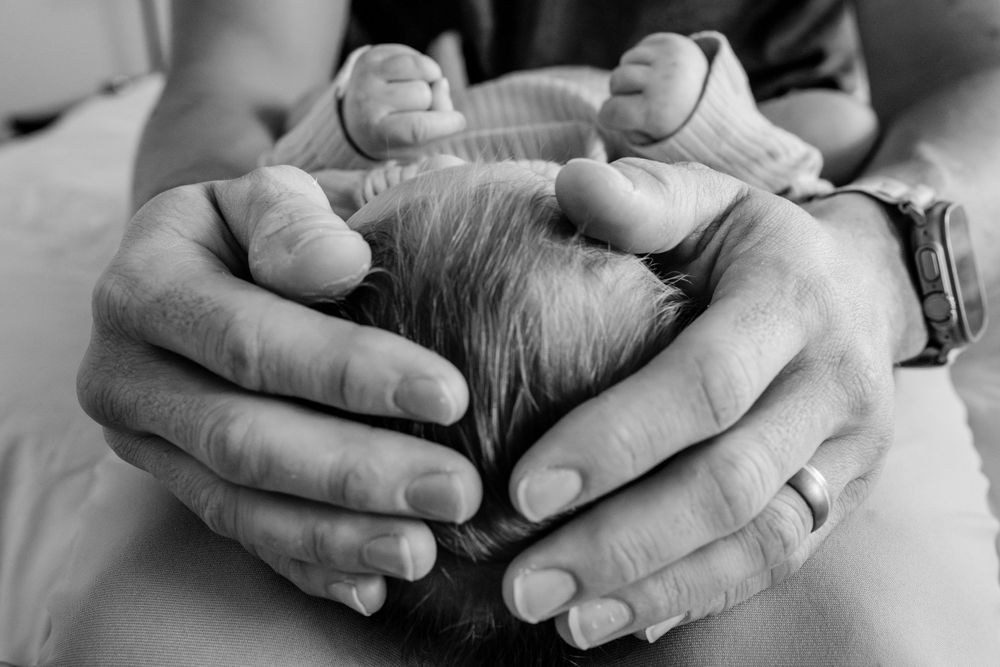 Gentle hands cradle a newborn's head in an intimate black and white portrait.