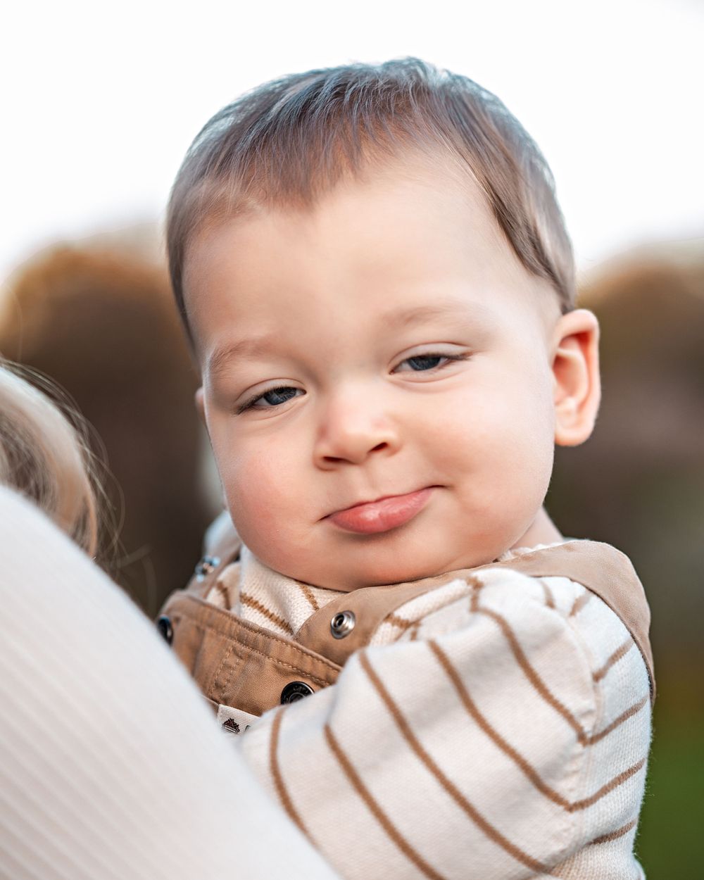 Adorable baby wearing a striped onesie smiles with rosy cheeks while being held outdoors.