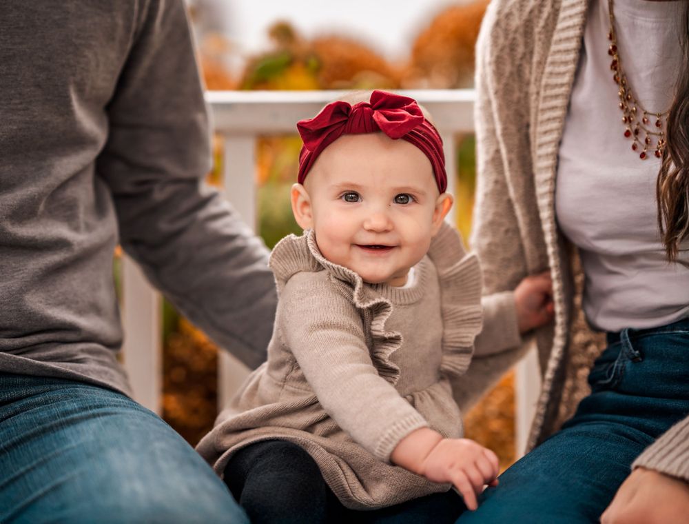 Baby in a tan sweater and red headband sits between two adults on a porch during autumn.