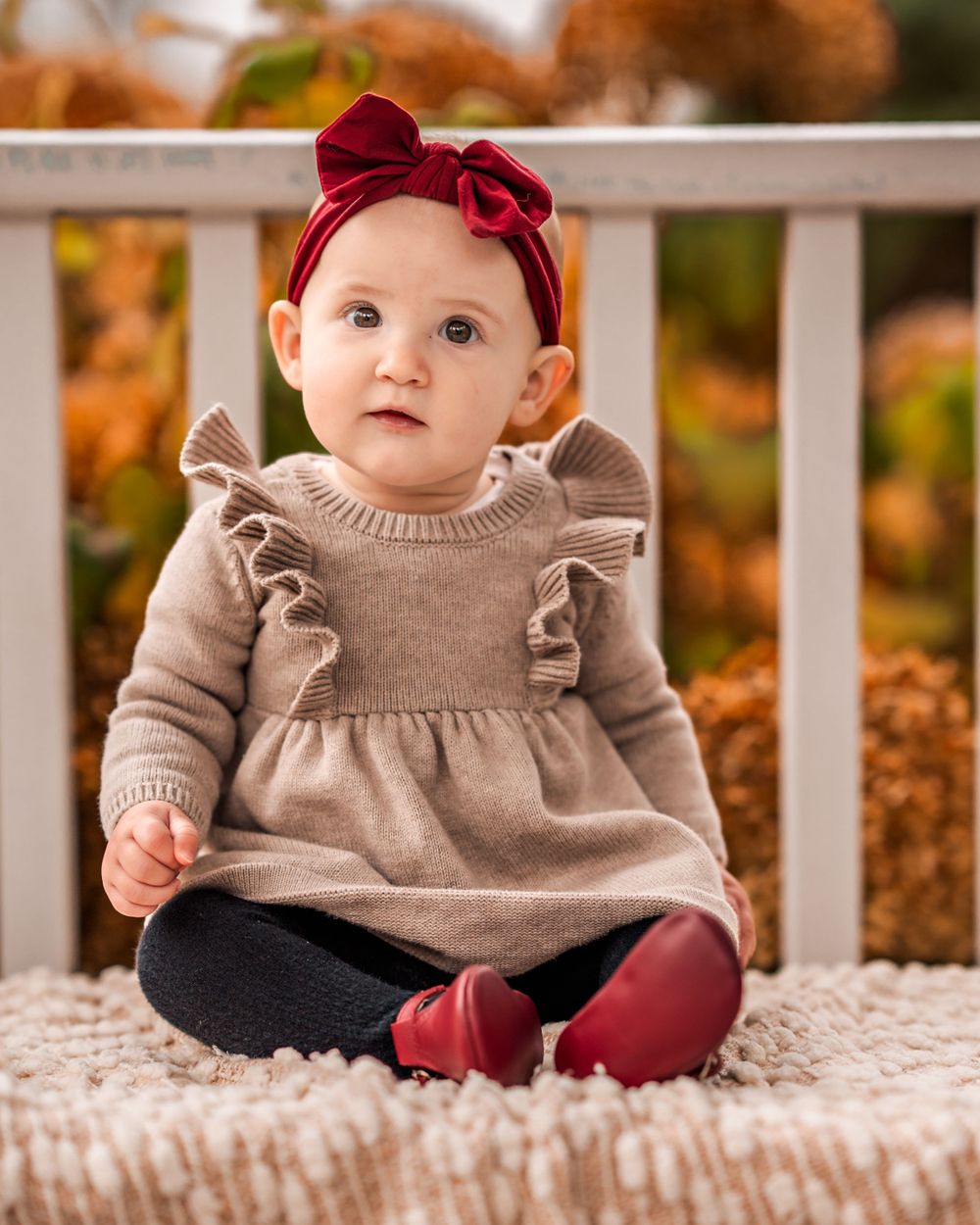 A baby wearing a beige ruffle dress with red bow headband sits on a white blanket during autumn.