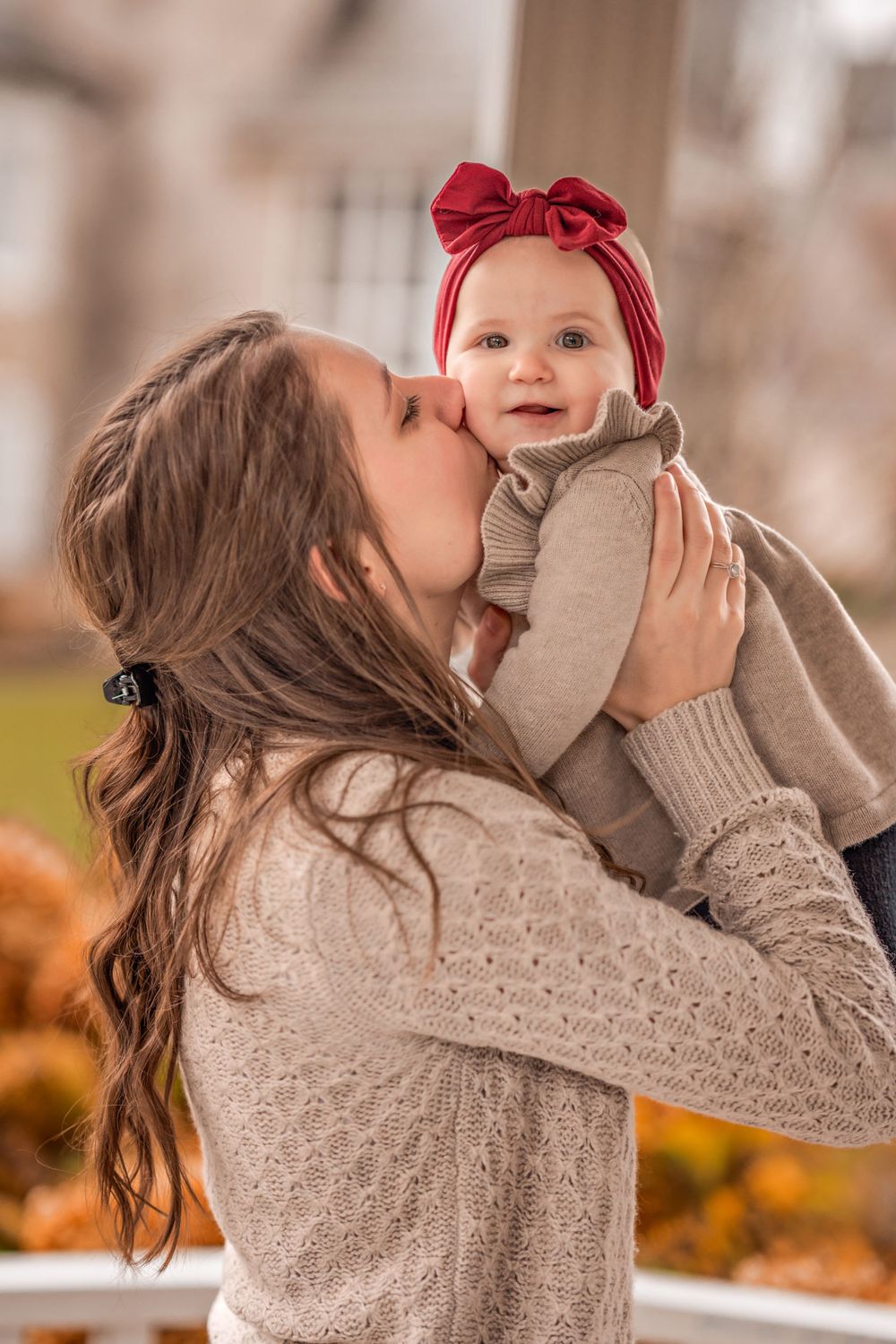 A loving moment captured in autumn as a parent lifts a baby wearing a red bow headband amid fallen leaves.