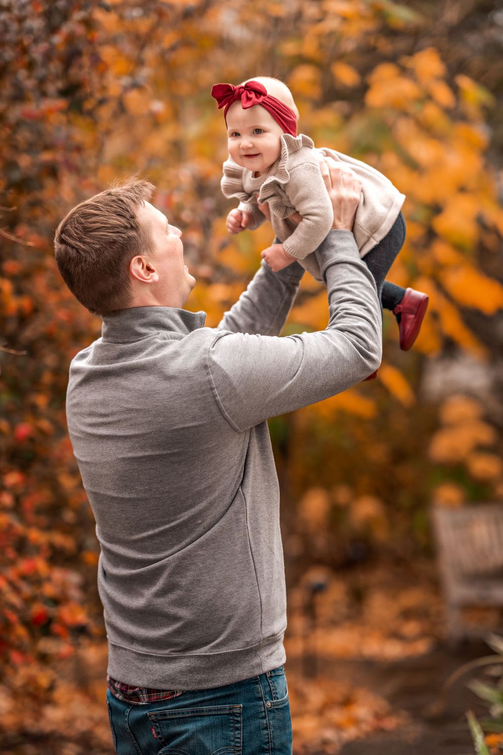 A person in a grey sweater lifts a small child wearing a red headband against a backdrop of autumn foliage.