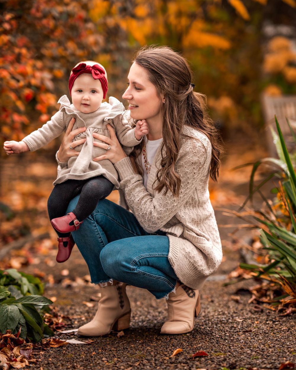 A parent and baby share a tender moment outdoors surrounded by vibrant autumn foliage on a path.