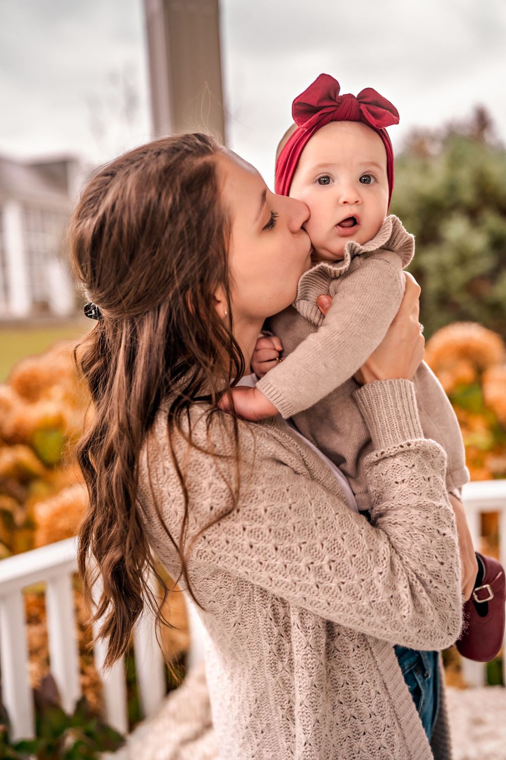 A tender moment captured in autumn as a parent kisses a baby wearing a red bow headband amid pumpkins and fall decor.