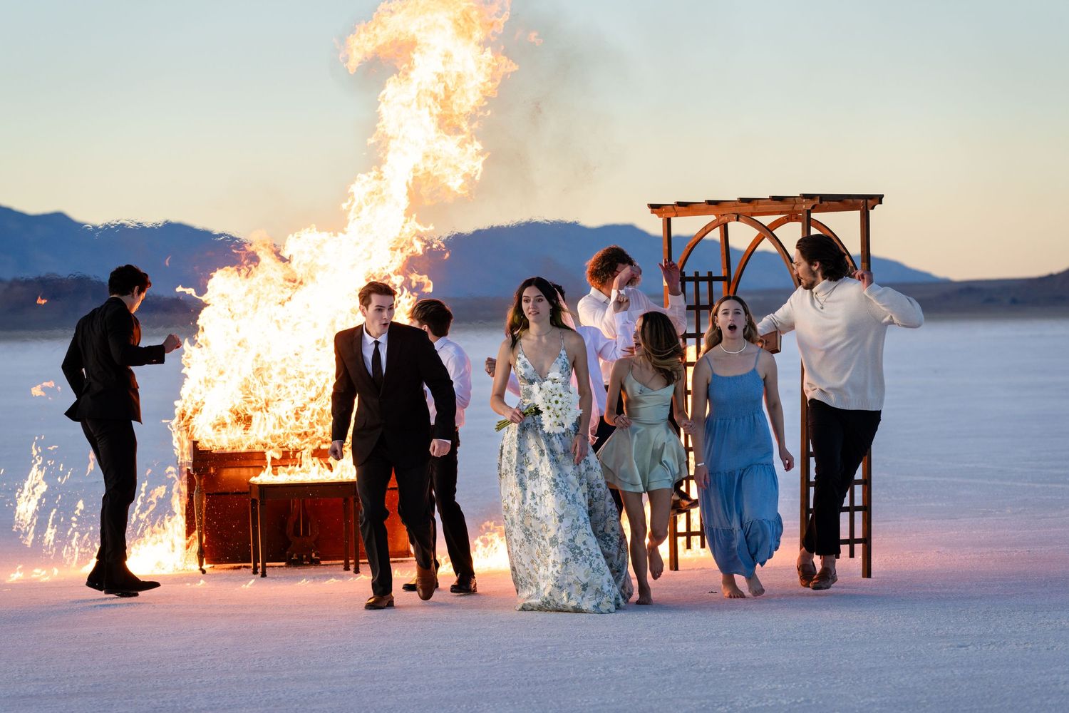 A wedding party gathered around a burning piano on salt flats at sunset.