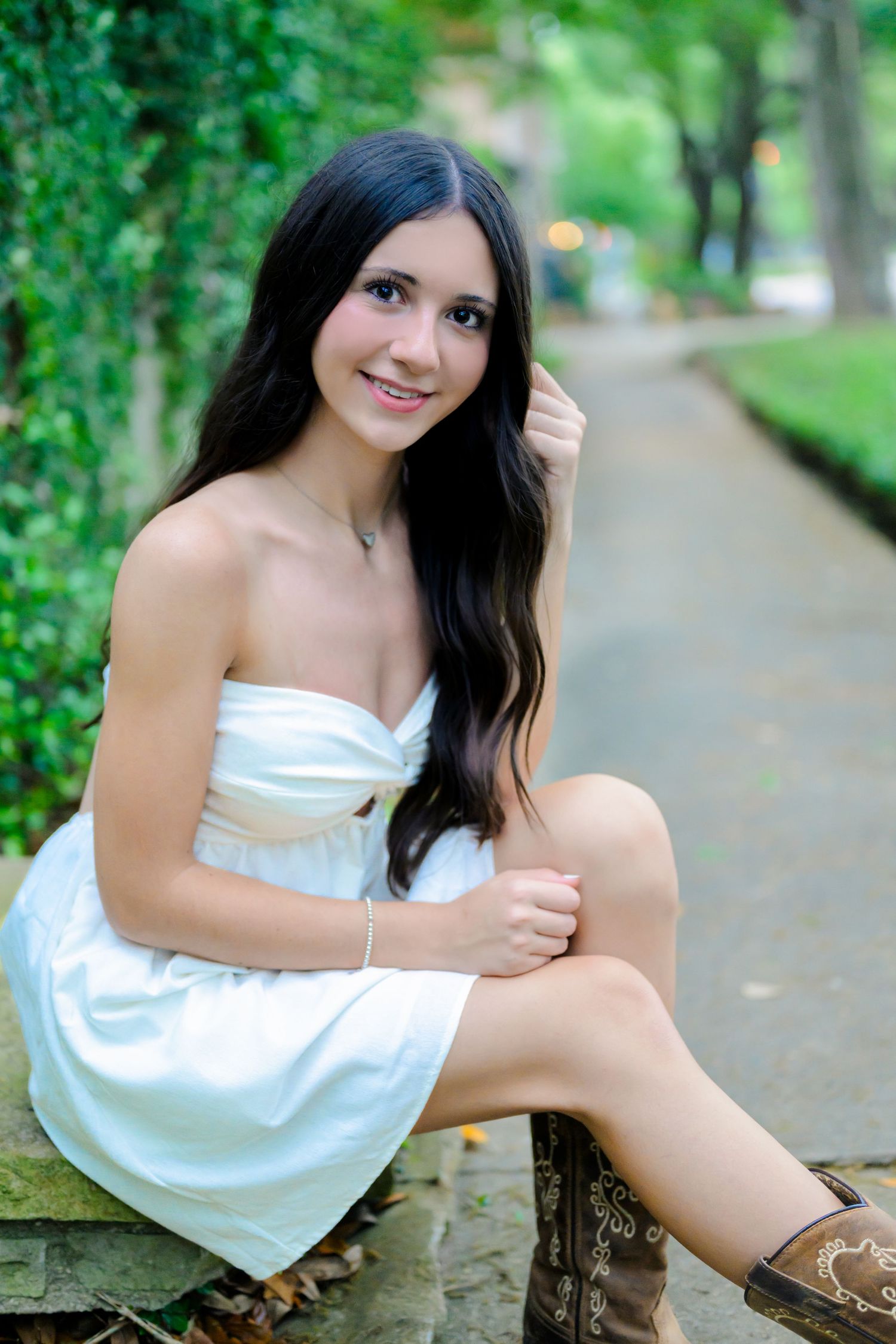 A person in a white strapless dress and brown cowboy boots sits on a rustic fence along a garden path.
