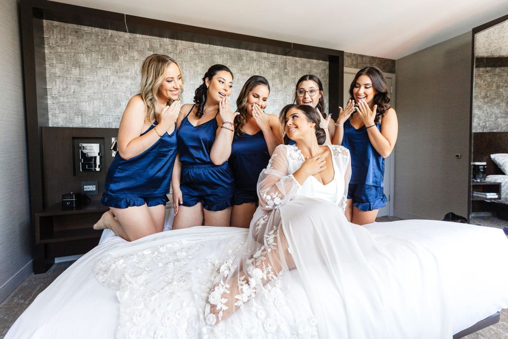 Group in matching blue robes and white lace wedding dress pose playfully on hotel room bed during pre-wedding preparations.