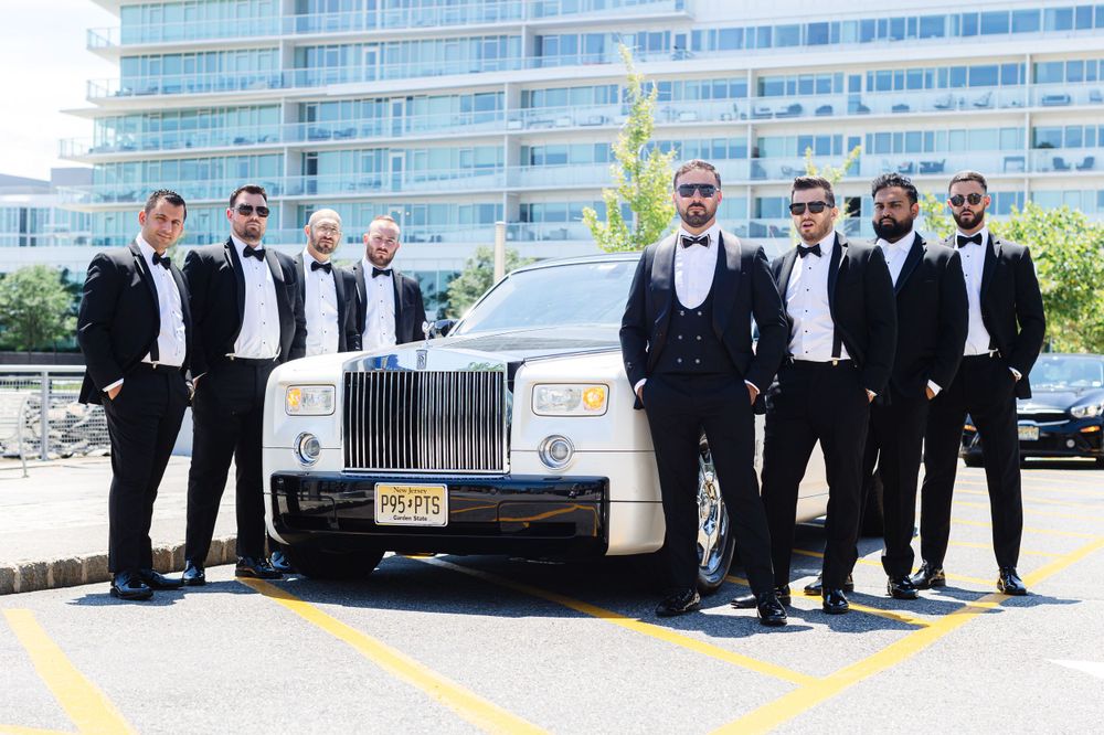 A group in formal black tuxedos poses next to a luxurious silver Rolls Royce vehicle outside a modern building.