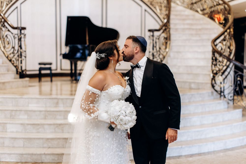 Wedding couple shares a romantic kiss on elegant marble staircase with grand piano and white floral bouquet.