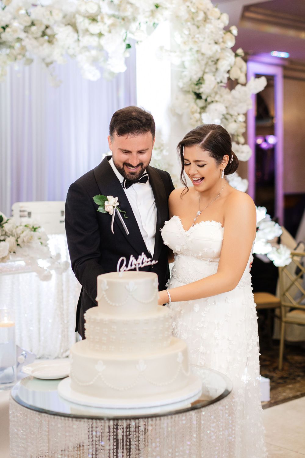 A newlywed couple shares a joyful moment while cutting their elegant white wedding cake decorated with flowers.