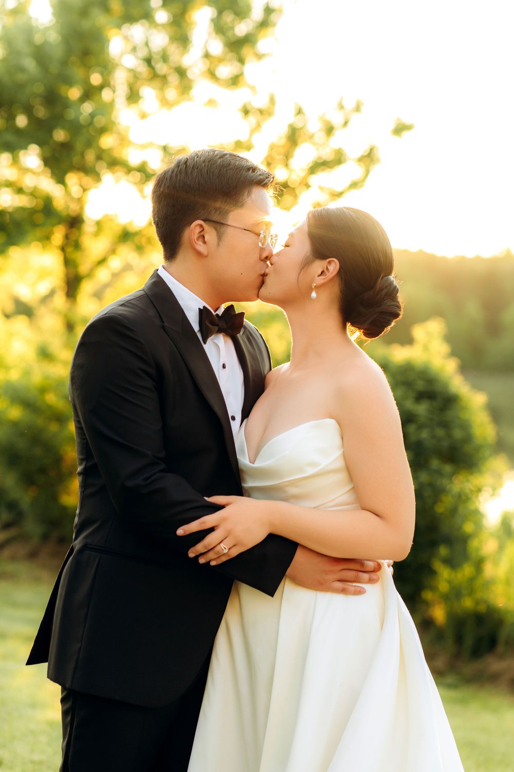 Bride and groom share a romantic kiss at sunset in formal wedding attire against a glowing natural backdrop.