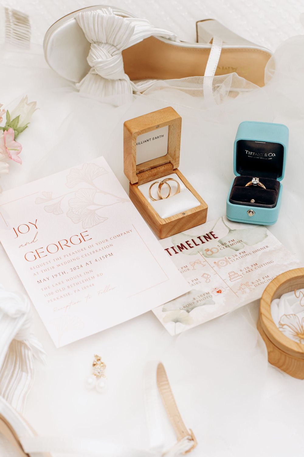 Wedding details flatlay with invitation, rings in boxes, timeline card and white heels against light background.