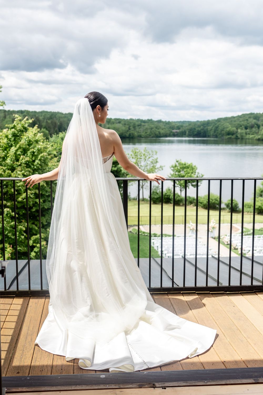 A flowing white wedding dress and veil seen from behind on a wood deck overlooking a serene lake view.