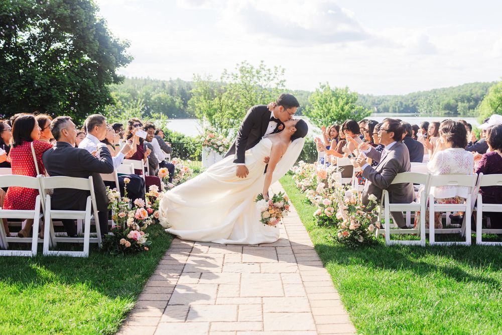 A romantic outdoor wedding ceremony on a stone patio surrounded by flowers with guests seated on white chairs.