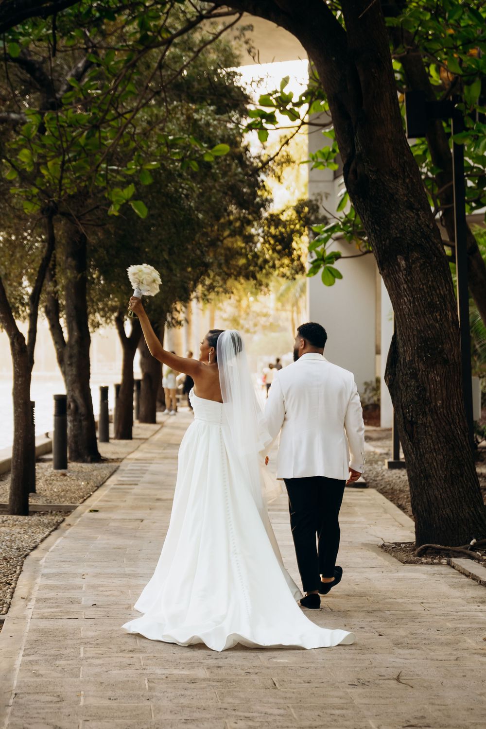 A couple in wedding attire walks down a tree-lined pathway lined with stone arches on their wedding day.