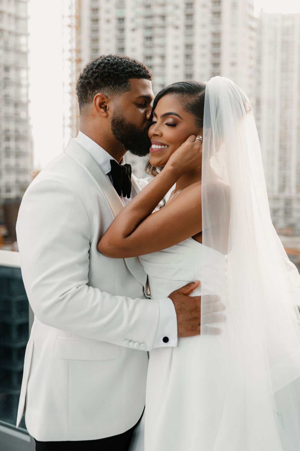 Tender wedding moment between newlyweds embracing in white attire against city backdrop with high-rise buildings.