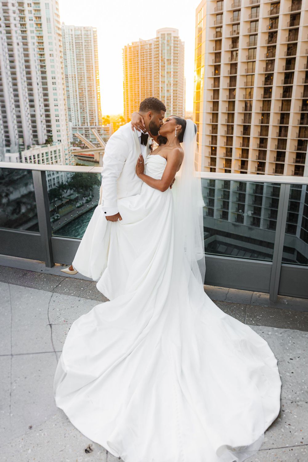 A romantic newlywed couple share an intimate moment on a high-rise balcony at sunset with city buildings in the background.