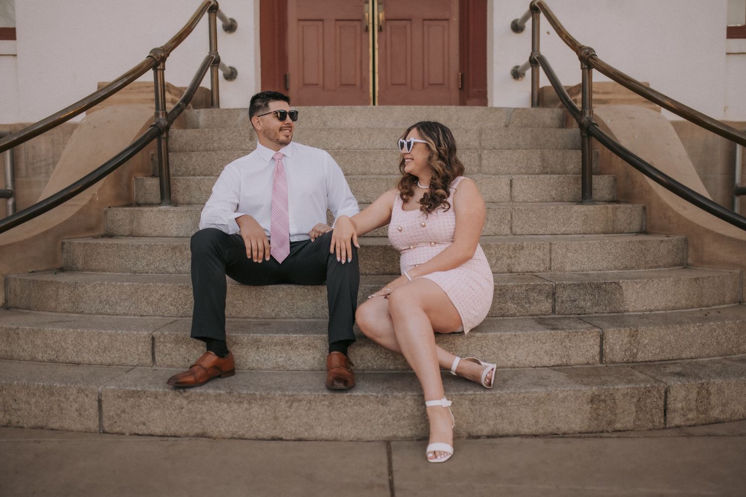 A couple sits together on stone steps in front of a door wearing pink and white formal attire in Downtown ABQ. Engagement photo style guide.