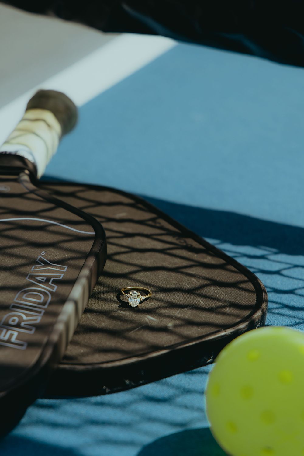 A diamond engagement ring resting on a Friday pickleball paddle next to a yellow ball on a blue court.