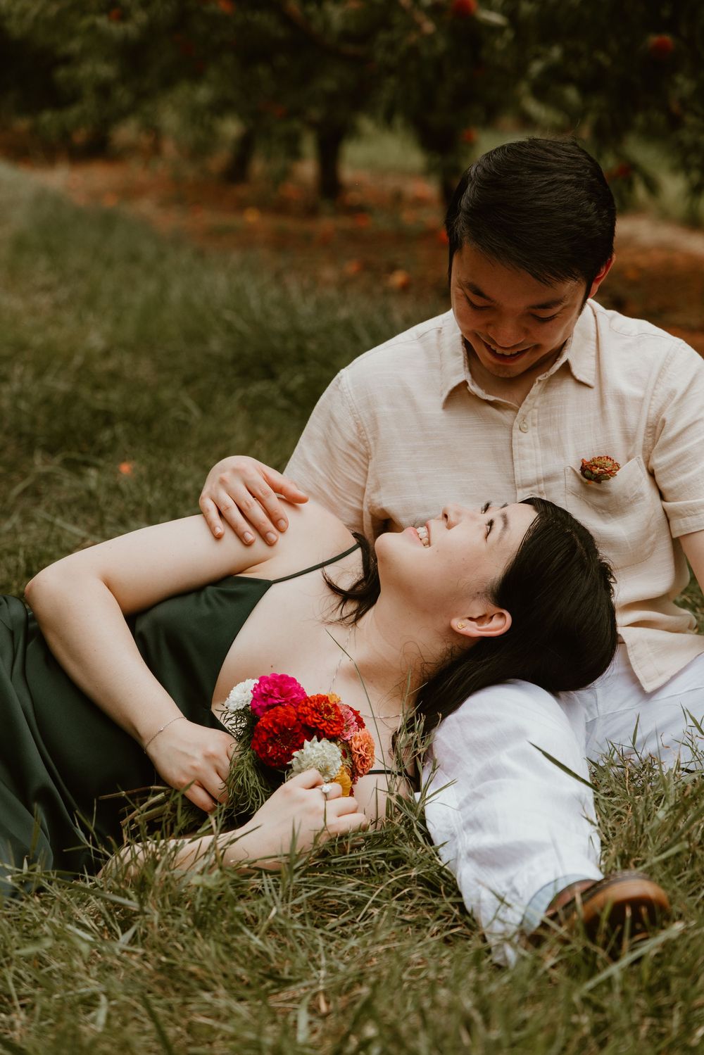 Couple relaxing in an orchard, woman lying in grass holding colorful flowers while man smiles down at her tenderly.