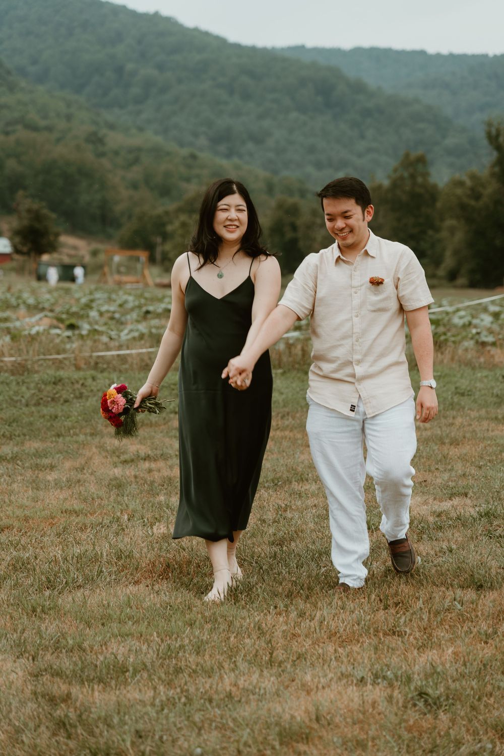 A couple walks hand-in-hand through a green field, she holds colorful flowers, mountains visible in the background.
