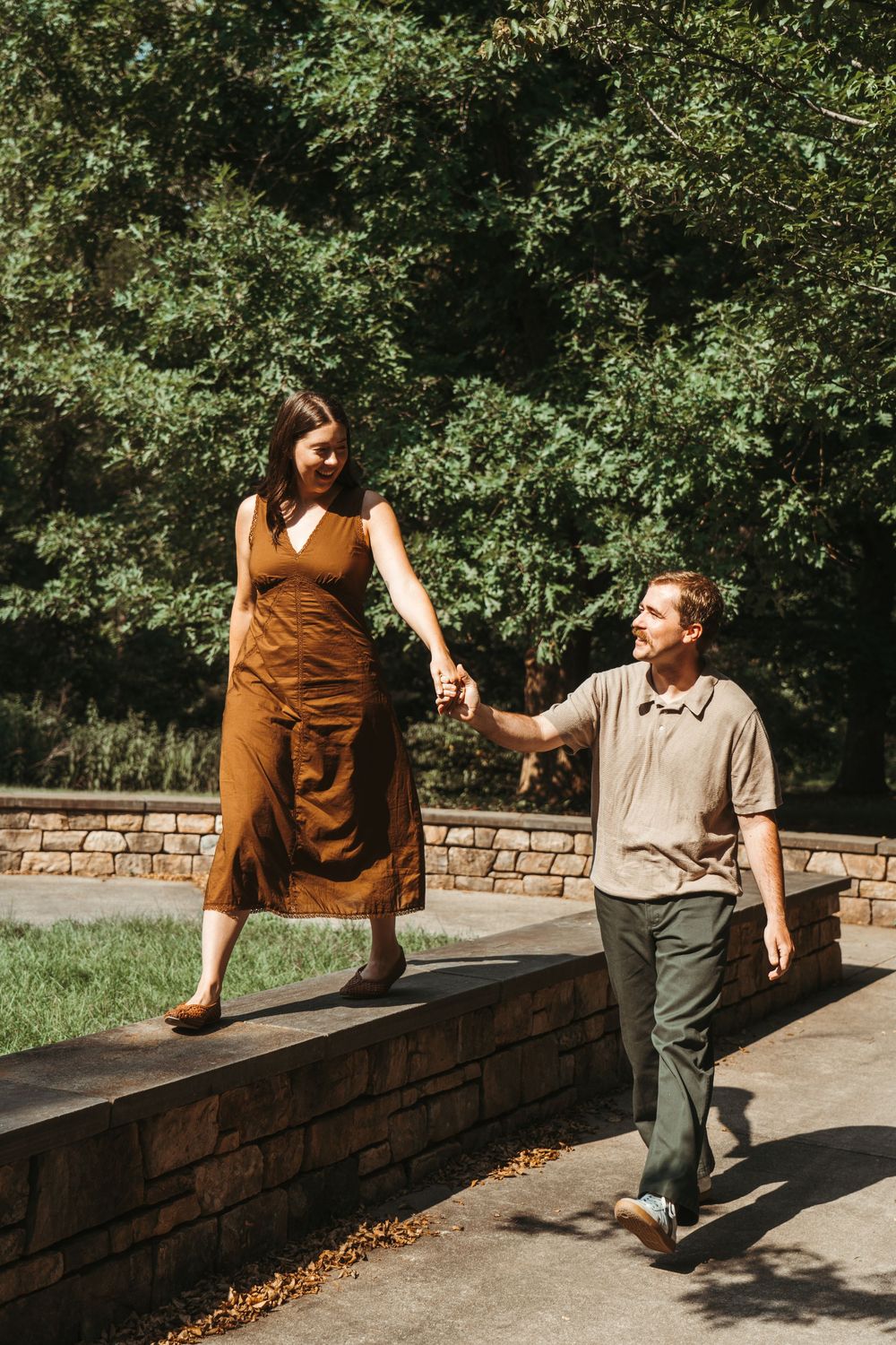 A couple holds hands while walking outdoors, woman in brown dress on stone wall, man in green pants, lush trees behind.