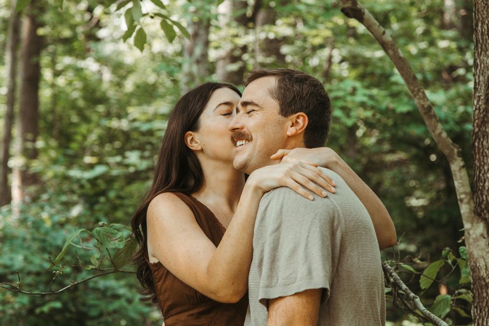 Couple sharing a romantic kiss outdoors surrounded by lush green trees in a forest setting.