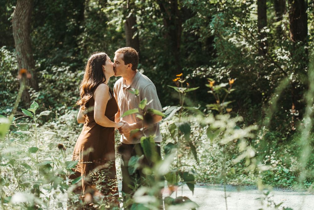 A couple shares a romantic kiss surrounded by lush green trees and wildflowers in a sunlit forest setting.