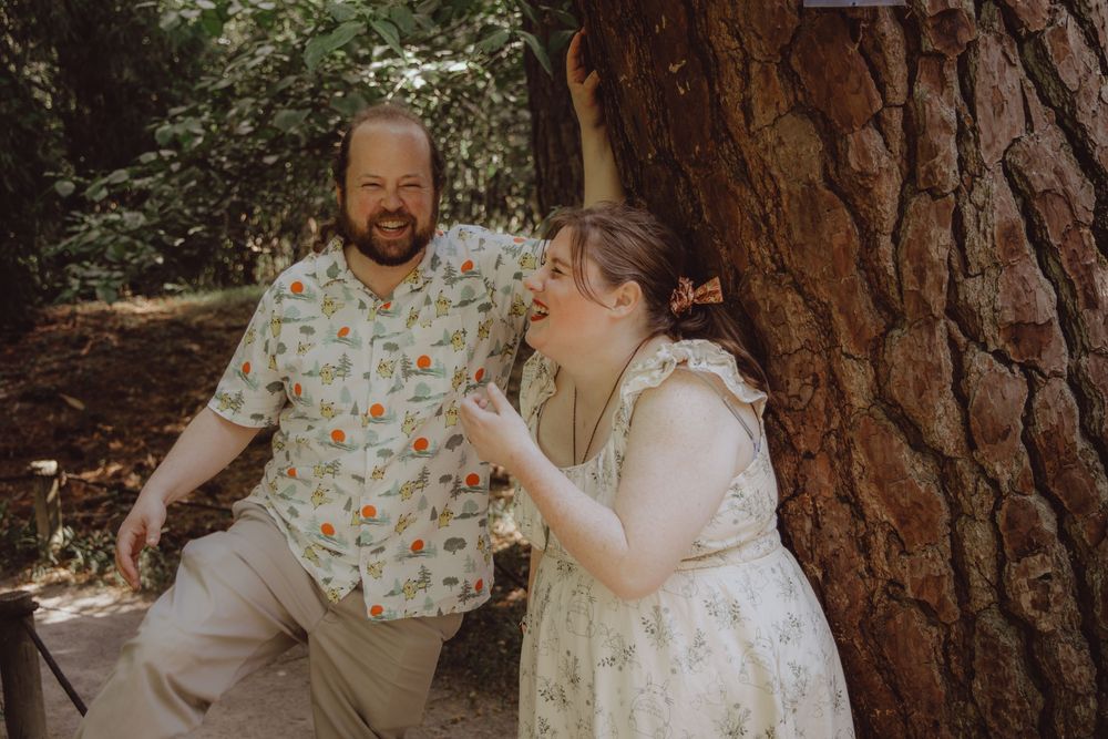 A laughing couple poses playfully by a large tree trunk in a sunlit forest, dressed in casual summer attire.