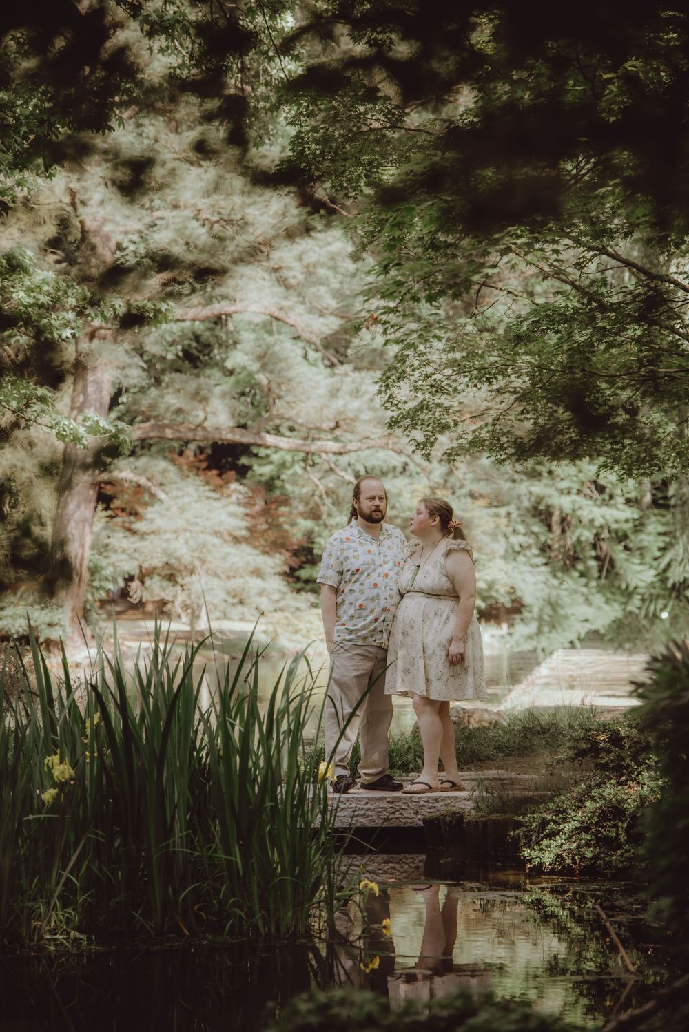 Couple standing on a wooden bridge surrounded by lush green trees and iris plants near a tranquil pond.