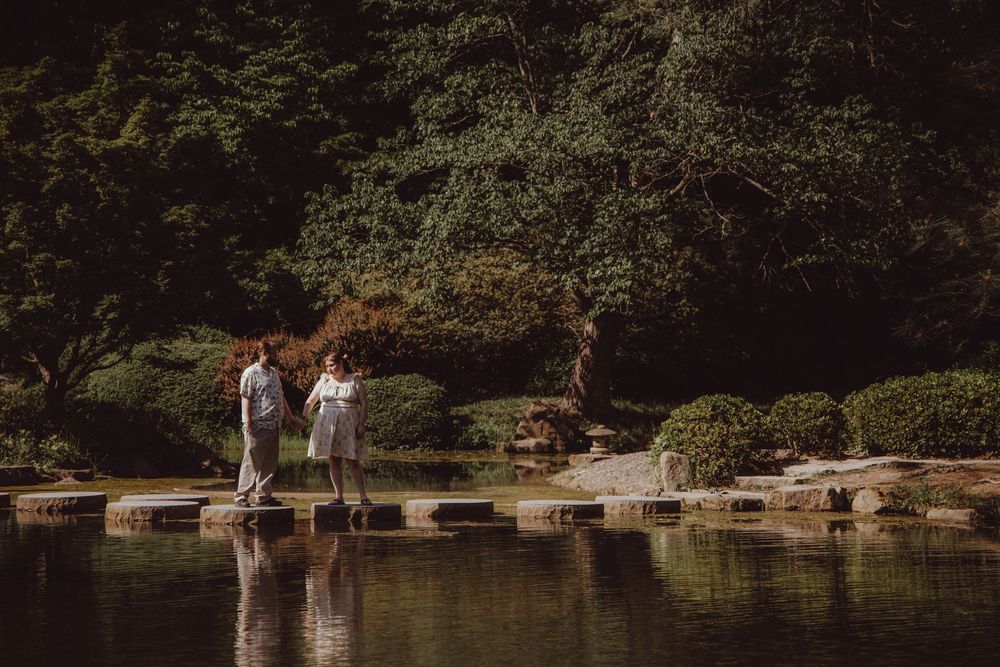 Couple standing on stepping stones over a tranquil pond in a lush Japanese garden.
