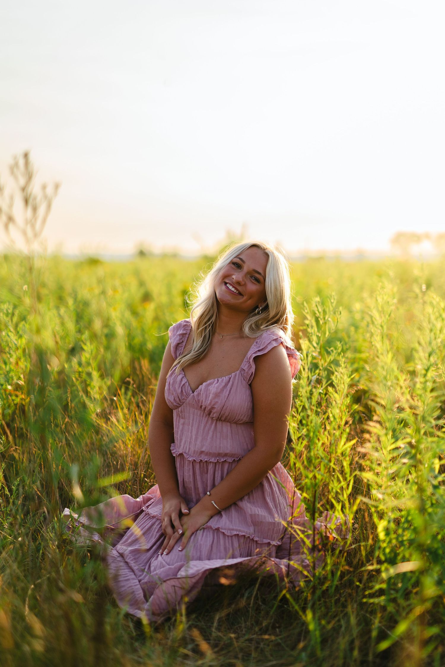 A person in a pink ruffled dress sits among tall grasses during golden hour sunset.