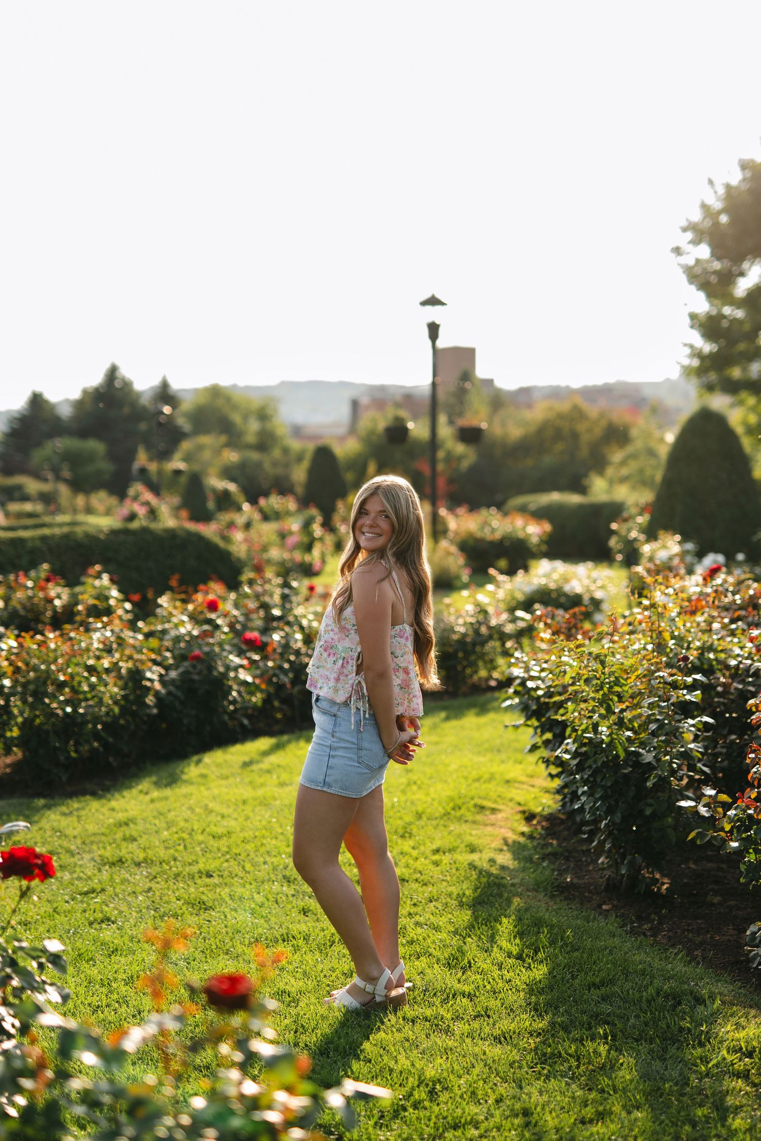 Smiling blonde in emerald green tank top poses outdoors against colorful summer foliage backdrop.