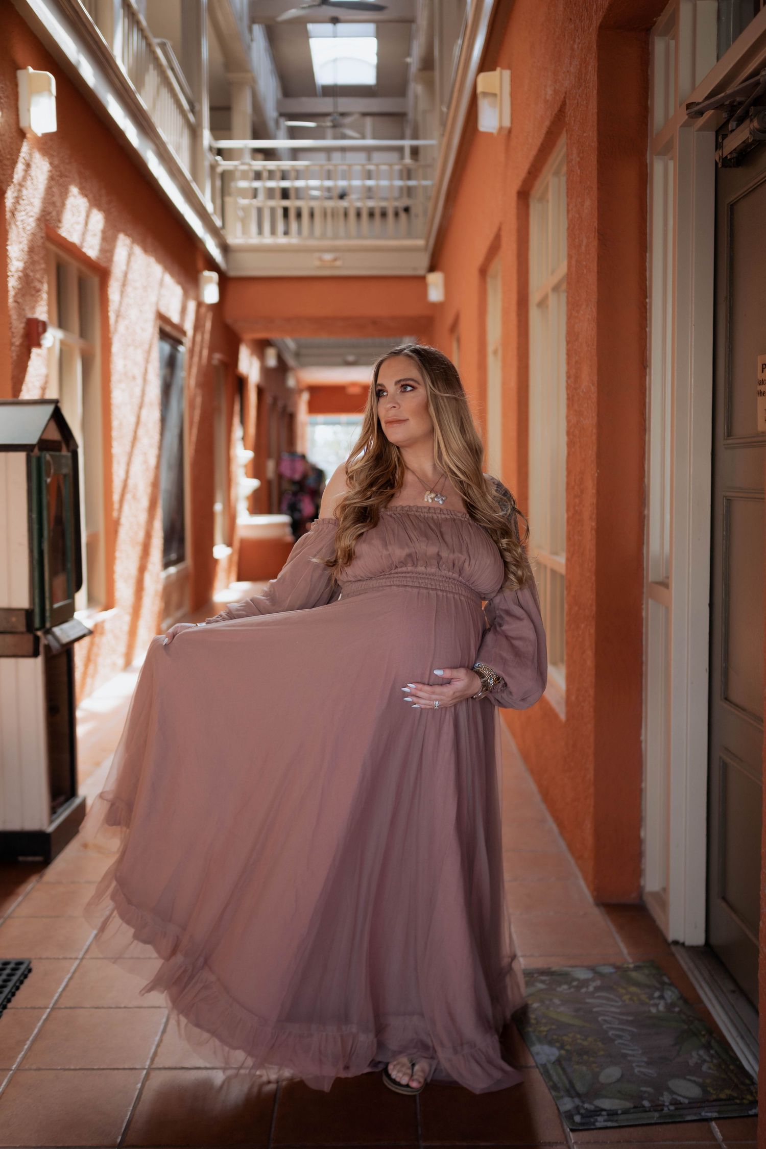 Pregnant woman in flowing dusty rose gown poses elegantly in sunlit coral-colored hallway.