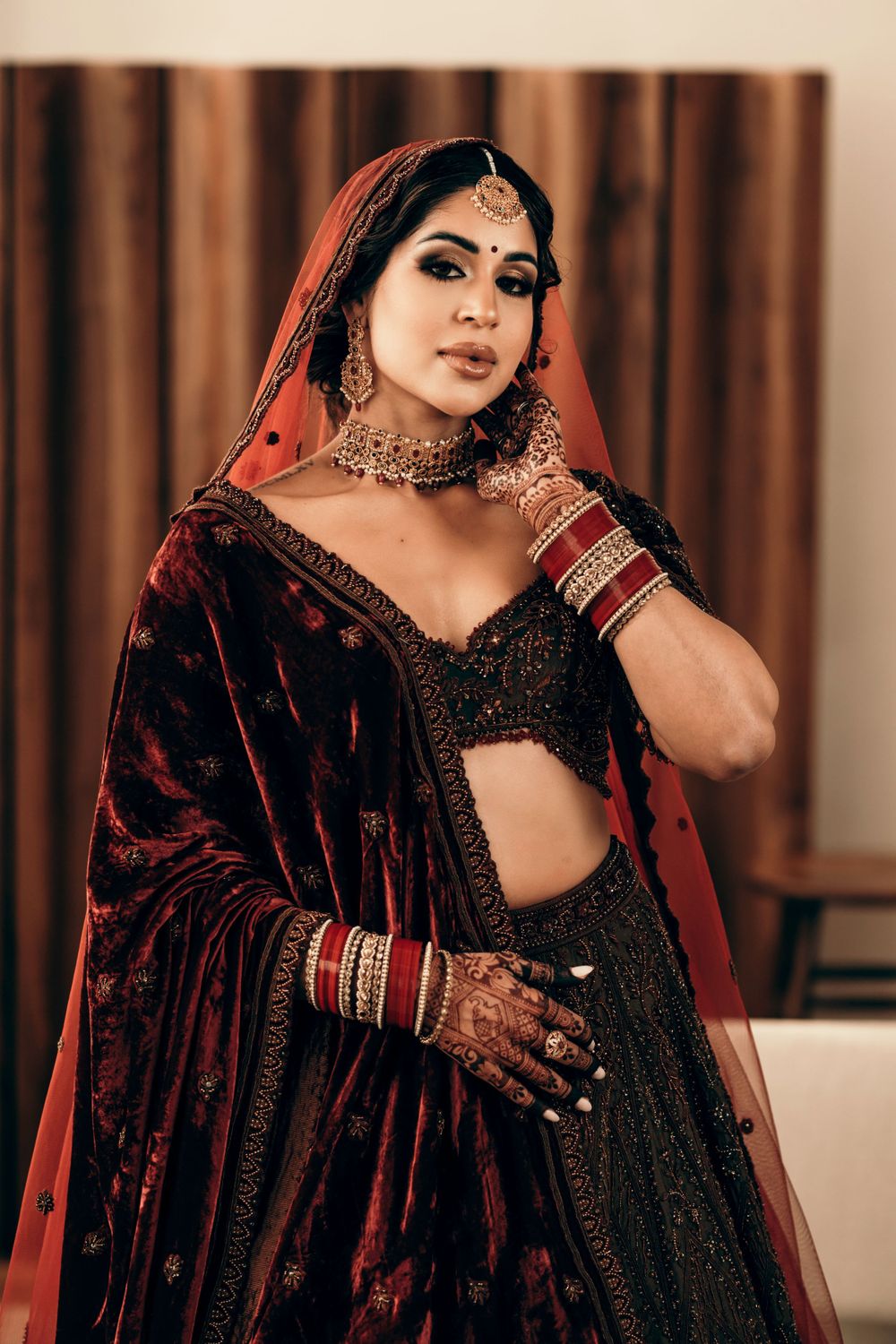 A woman in traditional South Asian bridal attire with dark velvet lehenga, ornate jewelry and henna-adorned hands poses elegantly.