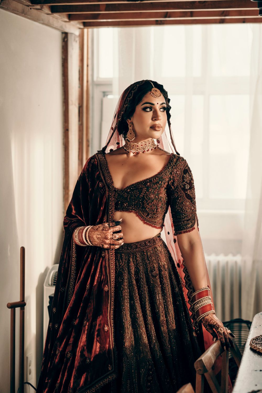 A bride in an elegant dark brown embellished lehenga with traditional Indian jewelry poses by a window.