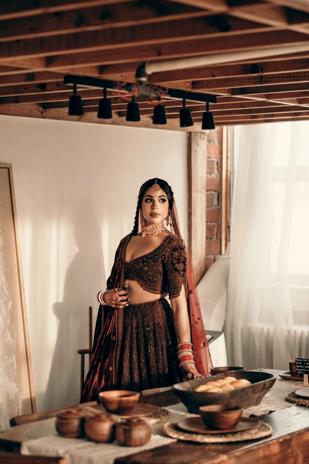 A person in traditional South Asian bridal attire stands in a kitchen setting with food displayed on a rustic wooden table.