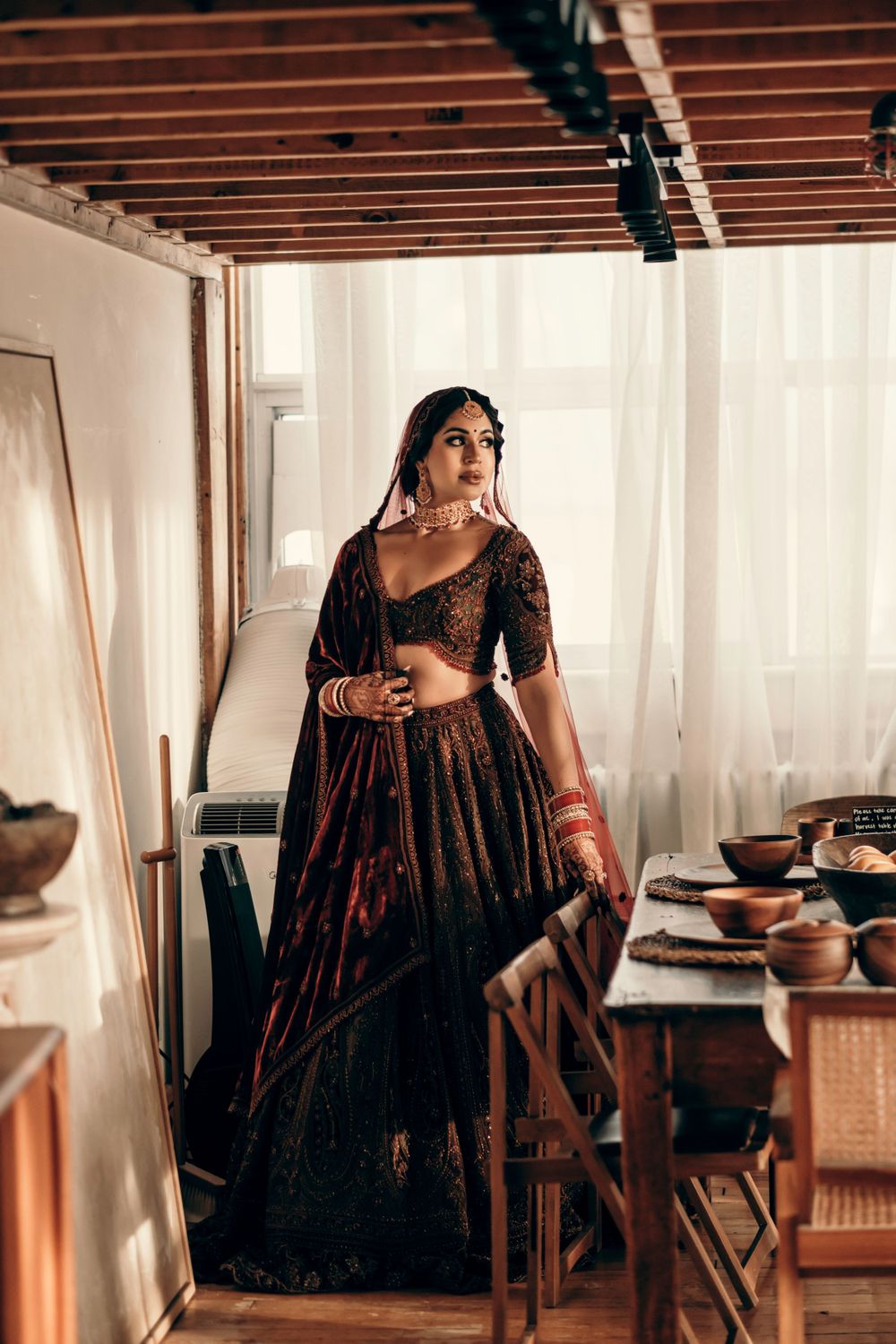 Woman wearing elegant dark velvet gown with detailed embroidery poses gracefully in rustic interior setting with wooden beams.