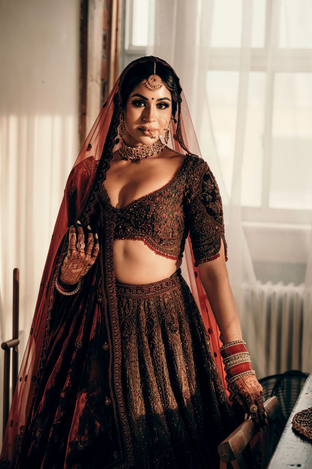 Traditional South Asian bride in dark embroidered lehenga and red dupatta poses elegantly by window light.