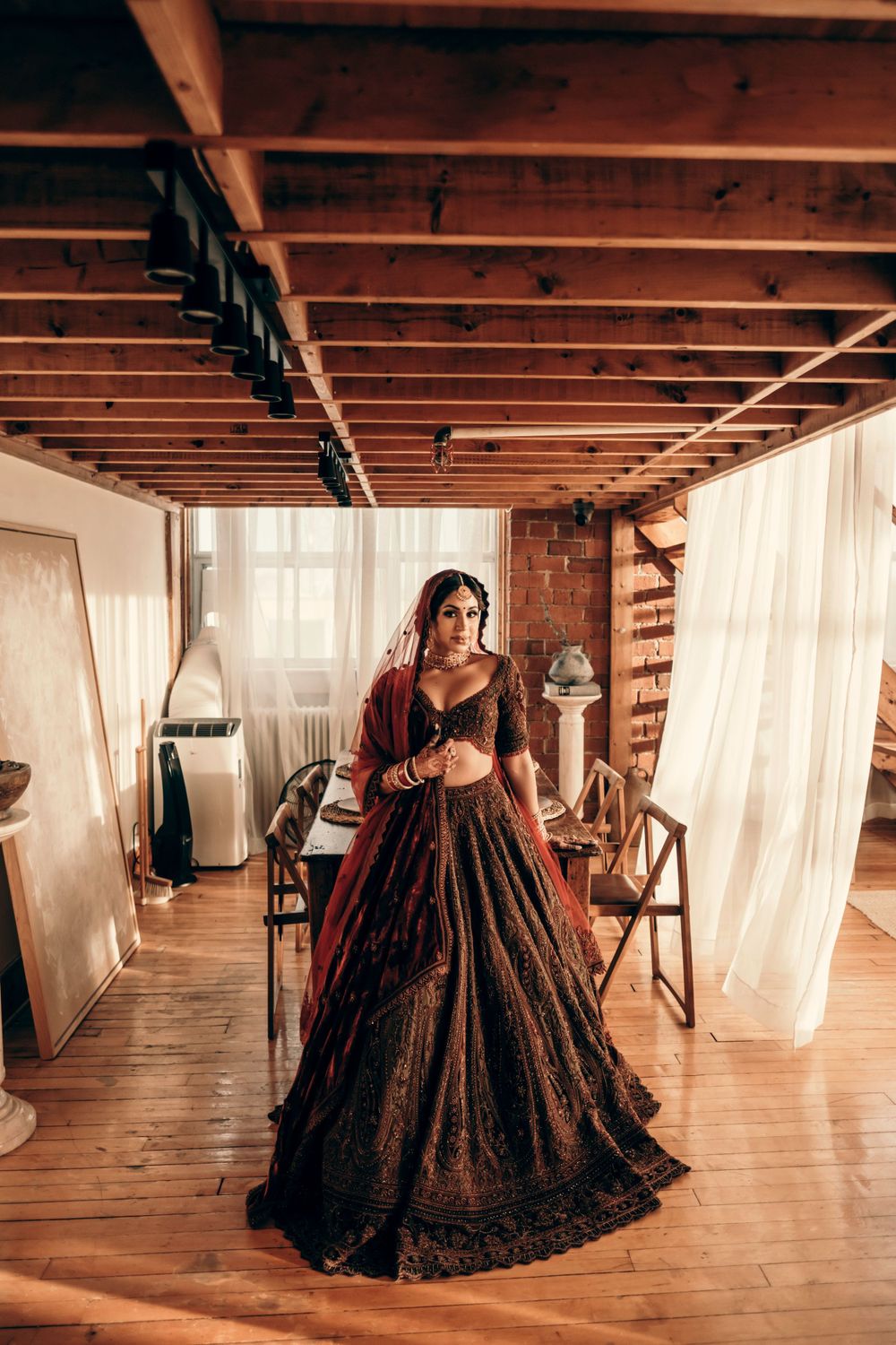 A woman in a burgundy ballgown stands in a rustic interior space with exposed wooden beams and flowing white curtains.