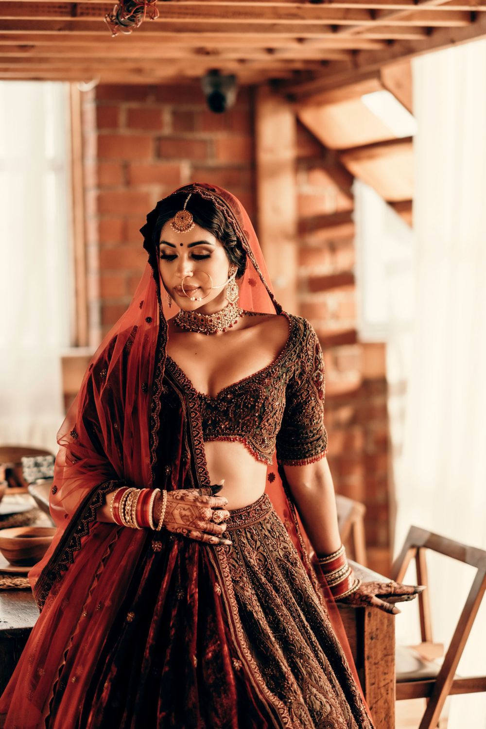 A bride poses in an ornate burgundy and gold lehenga with traditional Indian bridal jewelry and mehndi decorated hands.