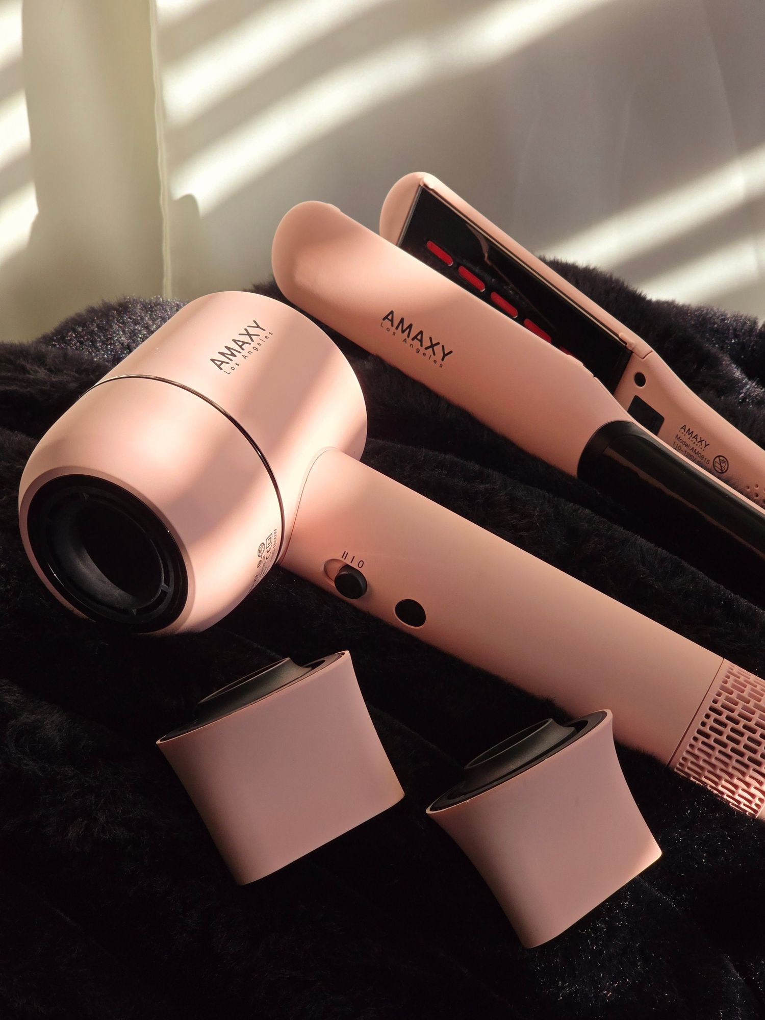 Rose gold hair styling tools including a dryer and flat iron with detachable attachments on a dark fabric surface.