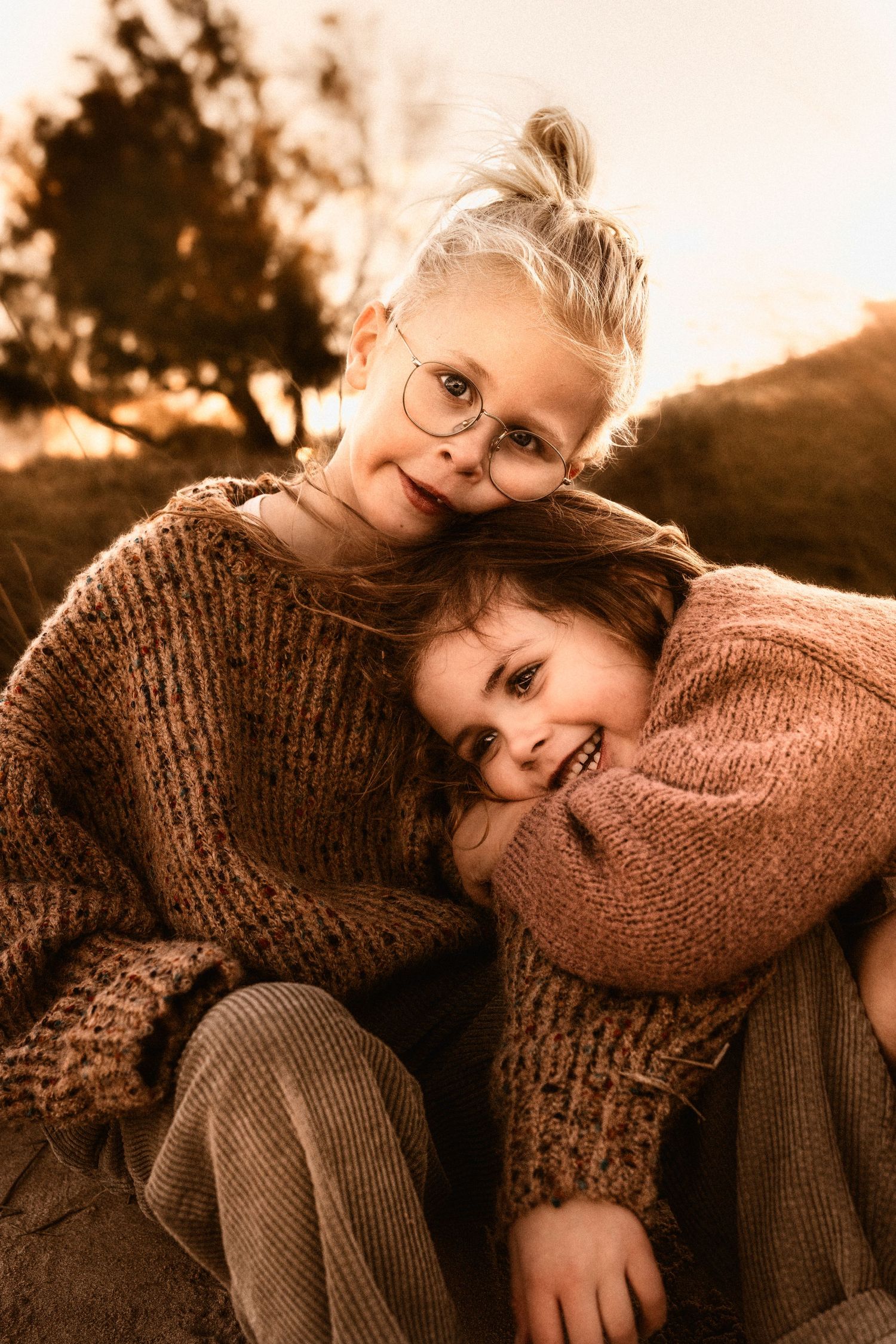 siblings in cozy brown sweaters in Denia share a warm moment together at sunset with a golden atmospheric glow, Javea Photographer