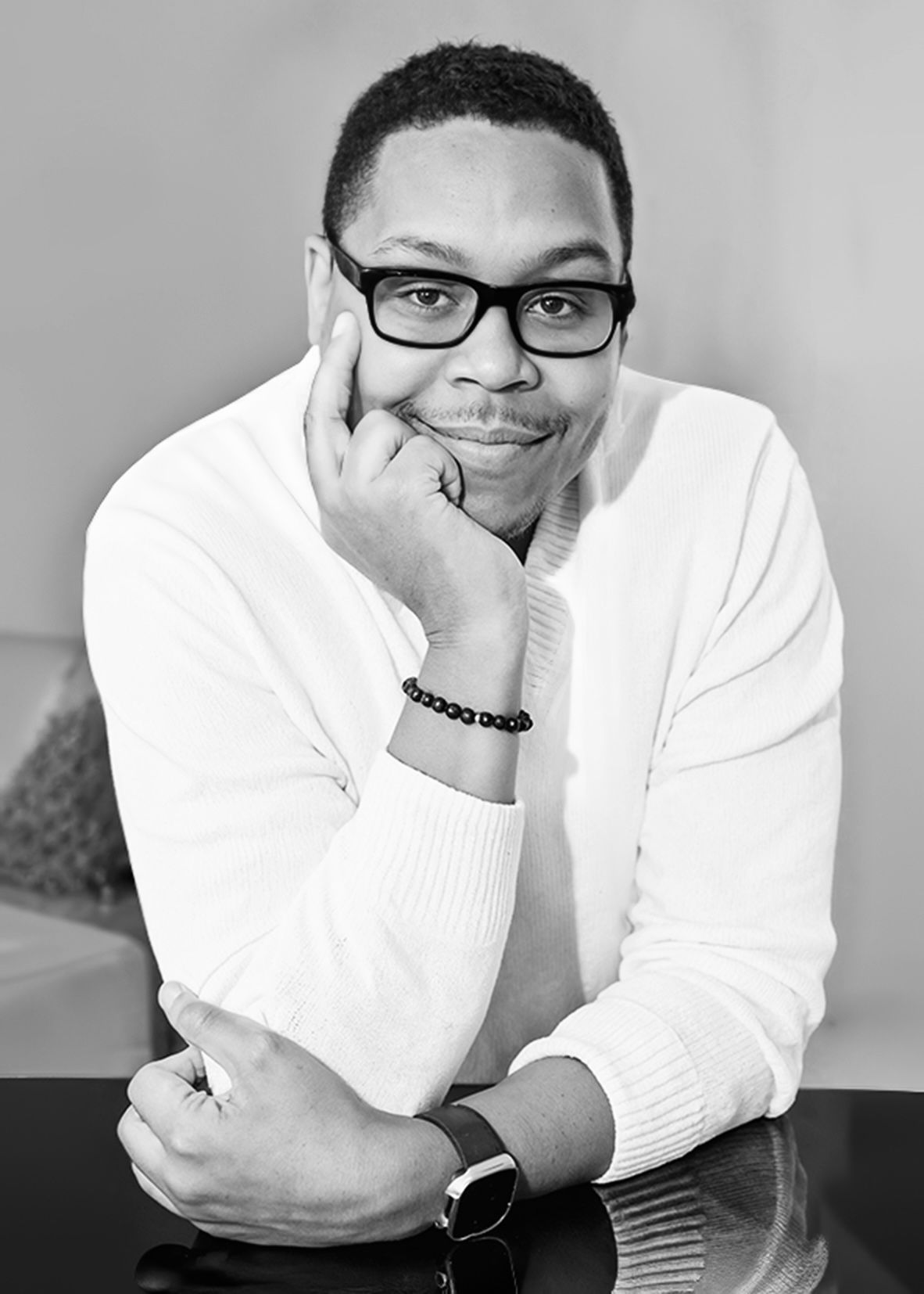 Black and white portrait of a smiling Black man in glasses and white sweater leaning on a surface.