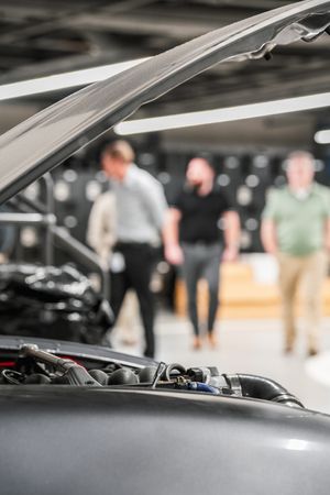 Close-up view of an open car hood with blurred figures examining a vehicle in the background of a showroom.