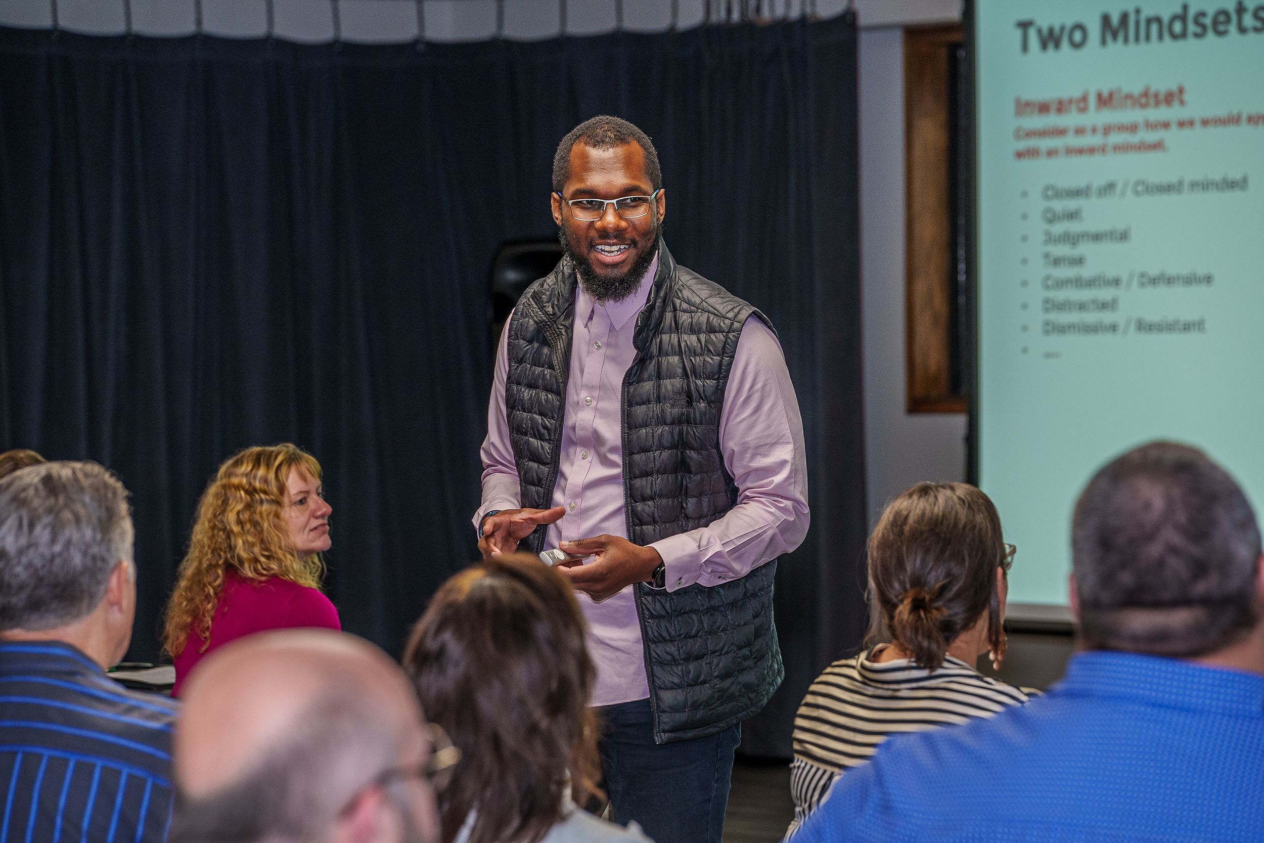 A presenter in a gray vest and light purple shirt addresses an audience during a business presentation.