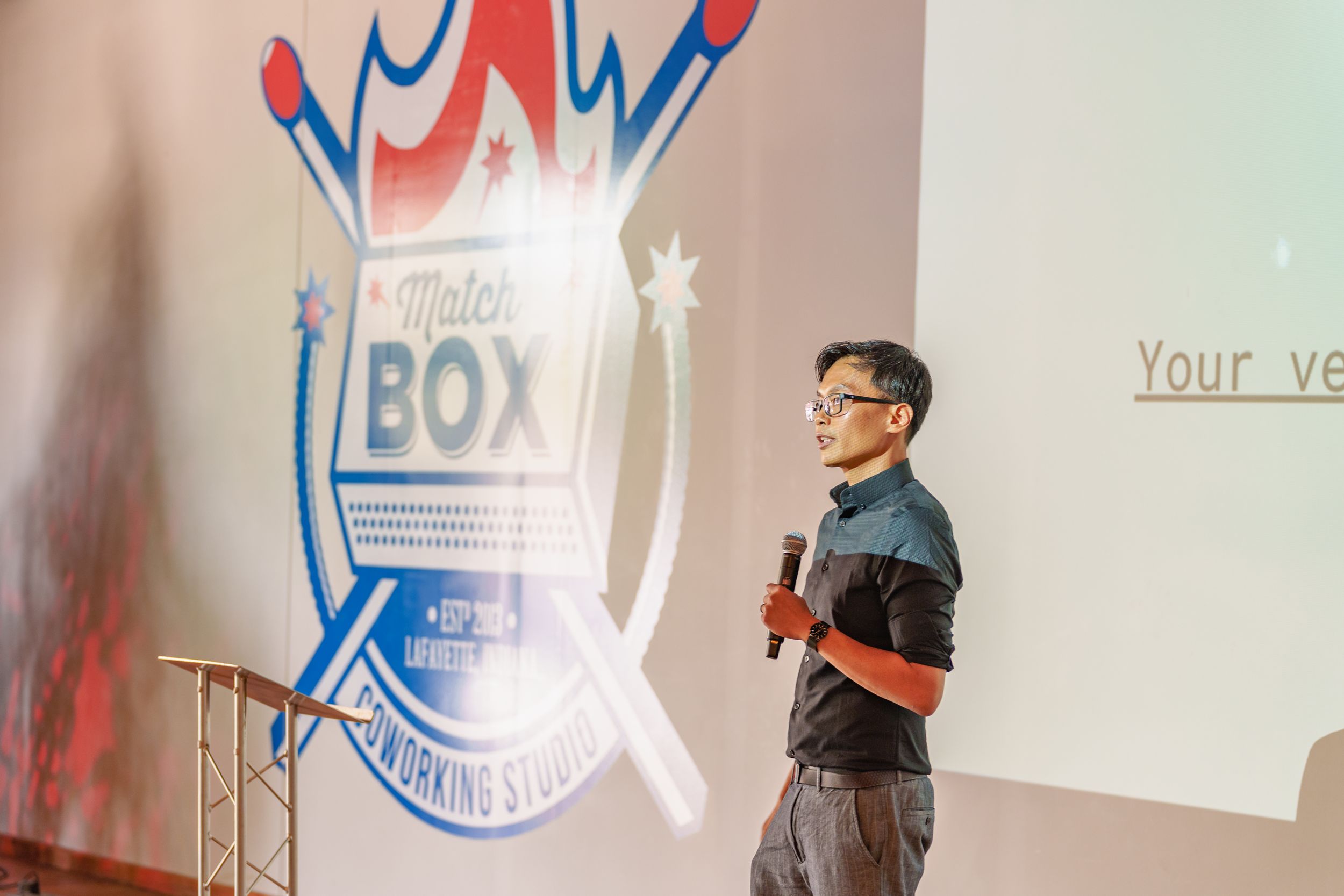 A speaker stands on stage giving a presentation with a projected screen and logo display in the background.