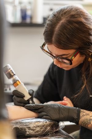 A tattoo artist wearing black gloves and glasses focuses intently while working with a machine in a studio setting.