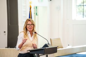 A speaker in a white blouse and glasses stands at a podium during a presentation in a bright conference room.